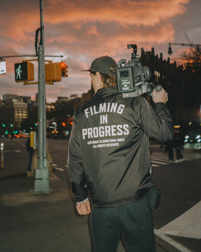 Person filming on a city street with 'Filming in Progress' jacket and sunset sky.